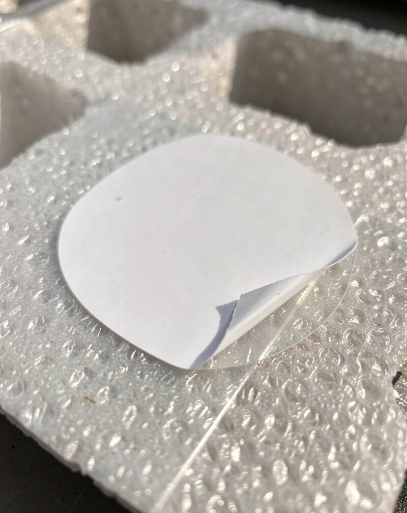 A blank white rectangular sticker half-peeled from wax backing paper lying on a car hood, adhesive sheen